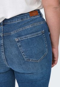 Jean en denim bleu délavé avec design à cinq poches, poche arrière cousue, et une étiquette de marque en cuir marron au-dessus de la taille.