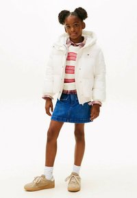 Biela prešívaná bunda s kapucňou, pod ňou ružovo-biely pruhovaný sveter, modrá rifľová sukňa a béžové tenisky s bielymi detailmi.