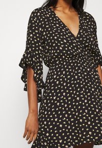 Schwarzes Blumenkleid mit tiefem V-Ausschnitt, kurzen Rüschensäumen und taillierter Passform. Es hat ein kleines Blumenmuster in Gelb und Pink.