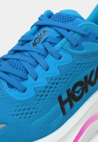 Baskets de sport bleues avec tige en maille, surcouches texturées, logo noir contrastant et semelle blanche avec une touche de rose. Les lacets sont plats et en synthétique.
