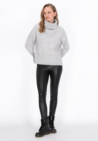 Maglione grigio a collo alto con lavorazione chunky, dotato di fessure laterali, abbinato a leggings neri in ecopelle e stivali neri alla caviglia con lacci.