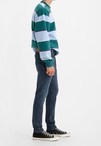Pull rayé vert, bleu clair et blanc avec un col haut, associé à un jean slim bleu foncé et des baskets noires.