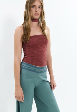 Jeune femme aux cheveux longs et blonds portant un haut bordeaux texturé sans bretelles, un collier assorti et un pantalon large bleu sarcelle avec une ceinture à motifs.