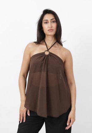 Vrouw met lang donker haar, gekleed in een bruine haltertop met centraal ringdetail en zwarte broek, staand voor een witte achtergrond.