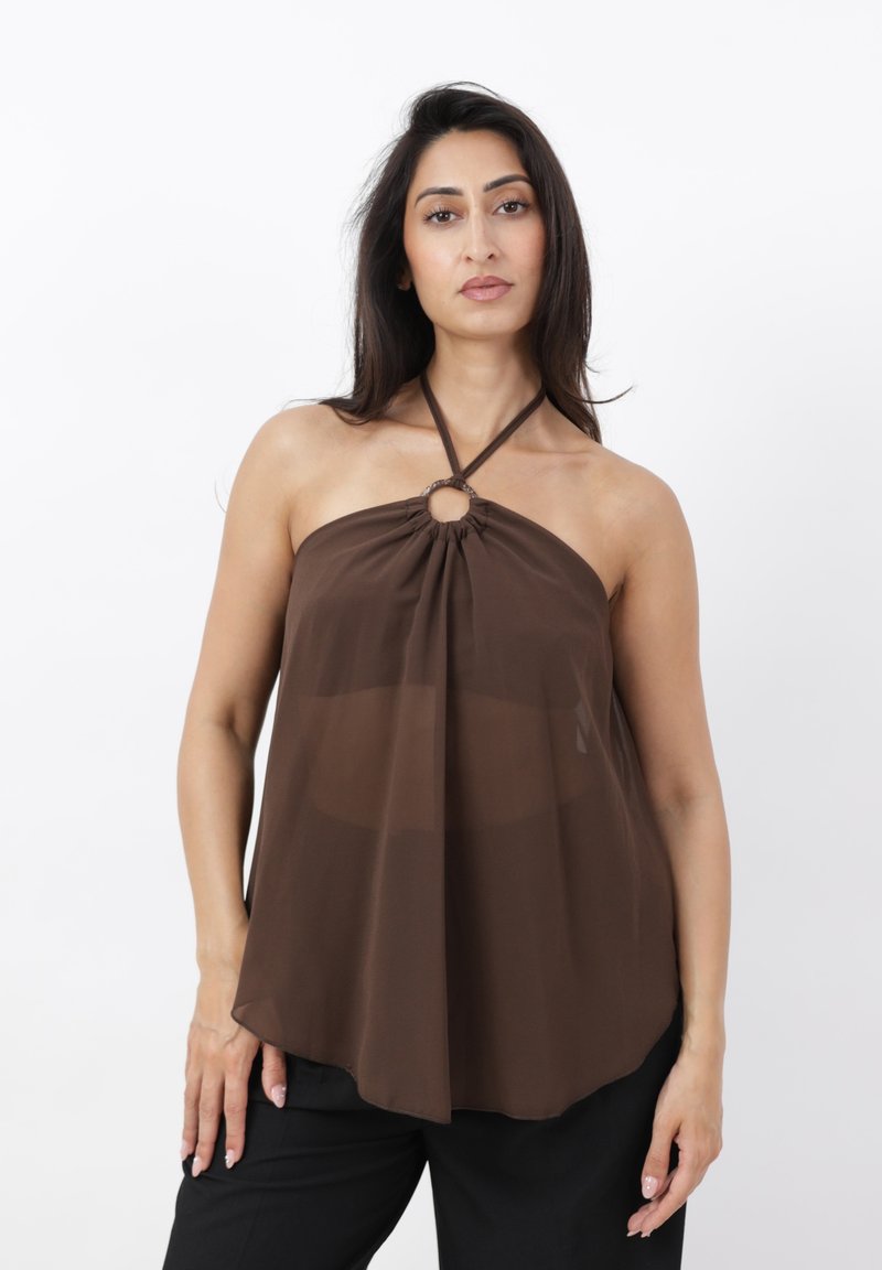 Mujer con cabello largo y oscuro que lleva un top marrón de cuello halter con detalle de anillo central y pantalones negros, de pie frente a un fondo blanco.