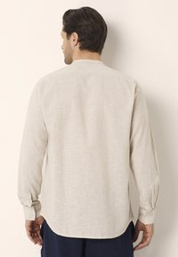 Camicia a maniche lunghe di un beige chiaro realizzata in tessuto texture. Presenta un colletto con bottoni e polsini bottonati, con un orlo leggermente curvato.
