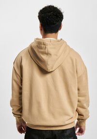 Beige kapucnis pulóver laza szabással, bordás mandzsettákkal és texturált anyaggal; kenguruzsebet és egy látható logót tartalmaz az ujján.
