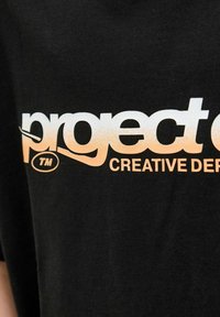 Černé bavlněné tričko s velkým bílým a oranžovým grafickým prvkem s nápisy "projekt" a "CREATIVE DEPT," má hladkou texturu.