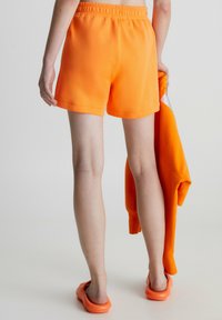 Oranssiset puuvillashortsit, joissa on joustava vyötärö, yhdistettynä matching-oranssiin collegepaitaan. Malli pitää collegepaitaa yhdessä kädessä.