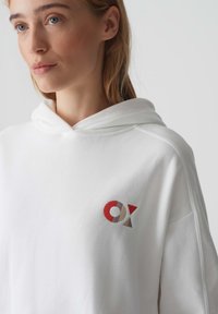 Felpa bianca con cappuccio regolabile e una texture morbida. Presenta un logo ricamato multicolore sul petto, che include rosso, marrone e grigio.