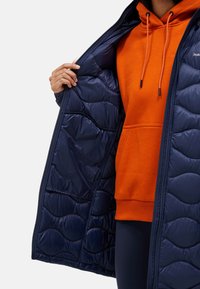 Marine pufferjack met een doorgestikte textuur, open geritst om een oranje hoodie eronder te onthullen. Het materiaal lijkt lichtgewicht en geïsoleerd.