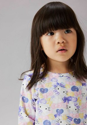 Jeune fille aux cheveux bruns et lisses, portant un t-shirt Hello Kitty pastel orné de cœurs et d'étoiles, sur un fond gris uni.