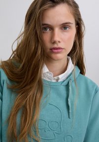 Young woman with long brown hair wearing a teal hoodie with a subtle skull design and a white collared shirt underneath.