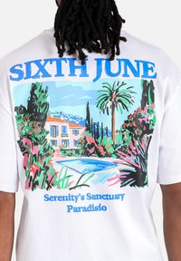 Witte t-shirt met een kleurrafische afbeelding op de achterkant, waarop een huis met een zwembad is afgebeeld, omringd door palmbomen en levendige bloemen. Bovenaan staat de tekst "SIXTH JUNE."