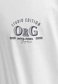 Bijela pamučna majica s crnim grafičkim printom koji sadrži natpise "STUDIO EDITION", "ORG", "JACK & JONES" i detalj potpisa. Glatka tekstura.