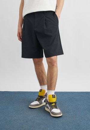 Persoon die zwarte knielange shorts, witte sokken en meerkleurige hoge sneakers draagt, staand op een blauw tapijt.