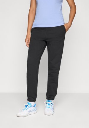 Person med svart joggebukse, hvite sokker, hvite og blå joggesko, og en lys blå ermeløs topp, som står med en hånd i lomma.