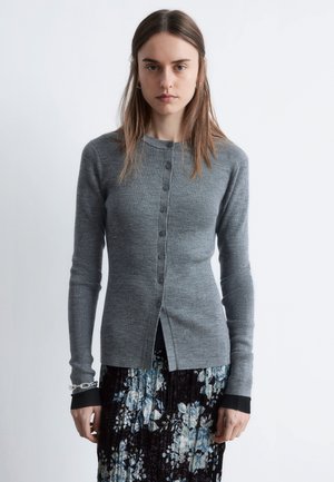 Graue gerippte Strickjacke mit Knopfleiste, langen Ärmeln und geteiltem Saum, kombiniert mit einem Rock mit floralem Muster.