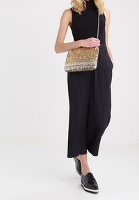 Kultainen ja hopeinen paljettikäsilaukku ketjuhihnalla, suorakulmainen muoto, ja monimutkainen kuvio, malli pukeutuneena mustiin vaatteisiin.