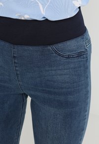 Lähikuva keskivartalosta, päällä sininen kukkakuosisen puseron, joka on työnnetty korkeavyötäröisiin sinisiin farkkuihin, joissa on leveä musta joustava vyötärönauha.