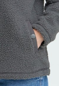 Hallus halli fliisist pullover, millel on tekstureeritud kanga, küljetaskud halli kangaga aktsentidega ja kontrastne nööpide detail.