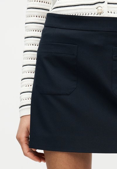 Κοντή ναυτική φούστα με ομαλή υφή, που διαθέτει δύο τσέπες πατίτες και καθαρό σχεδιασμό στη μέση, σε συνδυασμό με ριγέ λευκό πουλόβερ.