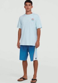 Young man standing wearing a light blue graphic T-shirt, blue gradient shorts with side stripe, and black flip-flops on a plain background.