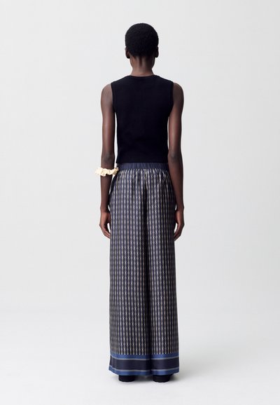 Una modelo está de pie de espaldas, vistiendo una blusa negra sin mangas y una falda larga con estampado, con un accesorio de color crema en el brazo.