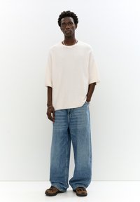 Maglione oversize beige chiaro a maniche corte, abbinato a jeans larghi in denim blu. Il modello indossa sneakers marroni scure con accenti a motivo.