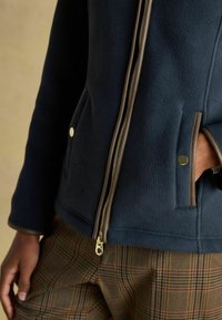 Osoba oblečená v námorníckej fleecovej bunde s hnedým lemovaním a zlatým zipsom, v kombinácii s hnedými kockovanými nohavicami, ruka v vrecku.