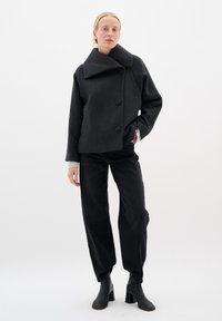Dunkelgraue oversized Wolljacke mit hohem Kragen und drei großen Knöpfen, kombiniert mit schwarzen weit geschnittenen Hosen und schwarzen Knöchelstiefeln.