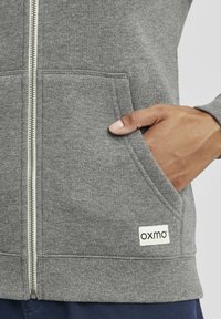 Gray zip-up sweatshirt with a textured fabric, side pockets, and a small white label featuring the brand "oxmo" at the hem.