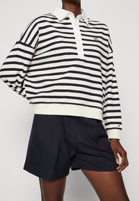 Osoba ubrana w biało-czarną w poziome pasy długą koszulę polo z guzikami i ciemnoniebieskie plisowane szorty.