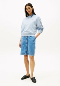 Felpa di cotone azzurro chiaro con colletto, abbinata a una gonna di jeans al ginocchio con chiusura a bottoni frontali e tasche laterali. Scarpe nere.