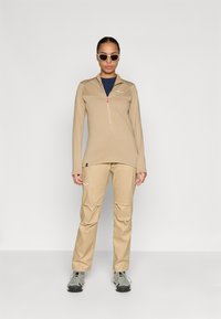 Μπεζ μπλούζα με μακριά μανίκια και φερμουάρ και αντίστοιχα παντελόνια, με τριγωνικό σχέδιο στην μπλούζα, φορεμένα με γκρι παπούτσια πεζοπορίας και γυαλιά ηλίου.