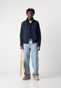 Veste coupe-vent marine avec col montant et fermeture éclair, associée à un jean bleu clair à jambes larges et des bottes beiges. Tient un sac fourre-tout beige.
