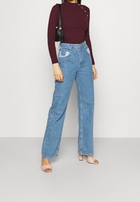 Maglietta a maniche lunghe a coste di un borgogna scuro con bottoni dorati, abbinata a jeans a gamba larga a vita alta di un blu chiaro con tasca a toppa. Tacchi beige.