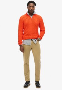 Pomarańczowy sweter z włóczki w splot, z zamkiem na szyi, nałożony na jasną koszulę w paski, w zestawie z khaki chinosami i czarnymi skórzanymi butami.