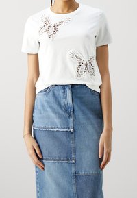 Bílý tričko s dvěma krajkovými aplikacemi ve tvaru motýlů. Kombinováno s modrou džínovou sukní, která vystavuje patchworkový design s roztrhanými okraji.