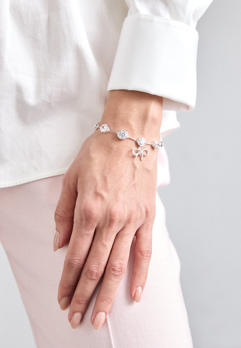 Ασημένια βραχιόλι με κυκλικά διαυγή πετράδια και λεπτομέρεια φιόγκου, με κομψό σχέδιο αλυσίδας και λείο υφή. Φορεμένο σε χέρι.