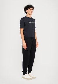 Svart bomulls t-skjorte med "Armani Exchange" logo, sammen med smale svarte joggebukser og hvite sneakers. Modellen står i profil.