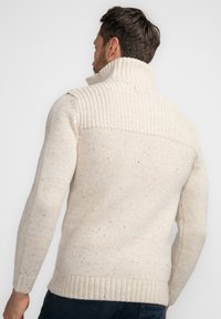 Cremfarbener Strickpullover mit hohem Kragen, geripptem Muster, gepunktetem Design und enganliegenden Ärmeln. Sichtbare Nähdetails an den Schultern.