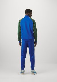 Track suit in blue and green, made of smooth fabric. Features a high collar, ribbed cuffs, and side stripes. Matching blue pants and sneakers.