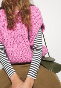 Chunky pletený růžový svetr s širokým výstřihem, na němž je navrstvená dlouhá černobílá proužkovaná košile s dlouhým rukávem, doplněný hnědými kalhotami a zelenou taškou.
