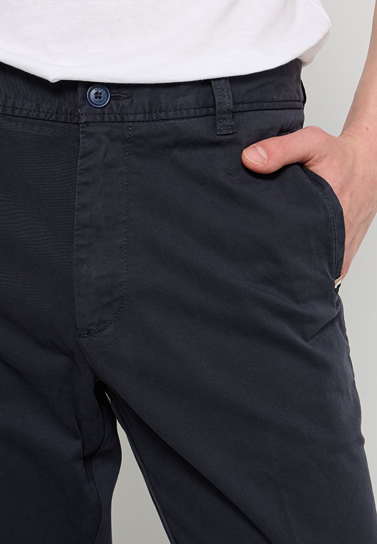 Nahaufnahme einer Person, die dunkle marineblaue Hosen mit einem blauen Knopf trägt, die Hand in der rechten Tasche und ein weißes Hemd, das hineingesteckt ist.