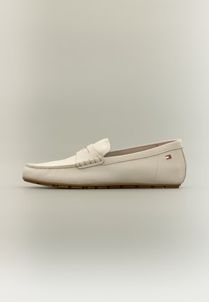 Beiger Lederloafer mit brauner Gummisohle, genähten Details und kleinem rechteckigem Logo an der Fersenseite vor neutralem Hintergrund.