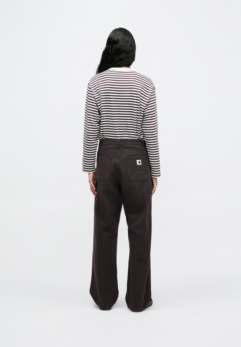 Persona con cabello largo y negro, vistiendo una camisa de manga larga a rayas y pantalones anchos oscuros, de pie de espaldas sobre un fondo blanco.