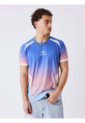 Man met krullend haar, gekleed in een blauw-roze verloop PXP t-shirt en lichte spijkerbroek, kijkt naar rechts met één hand in zijn zak.