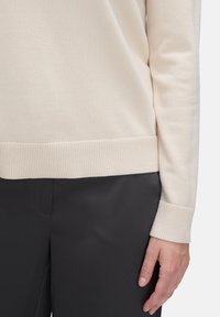 Persoon in een crèmekleurige geribbelde trui met boorden en zwarte broek, met de rechterhand naast zich op een witte achtergrond.