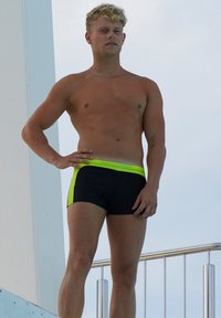 Schwarze Schwimmshorts mit einem neongrünen Akzentstreifen an den Seiten, gezeigt an einer Person, die auf einem Sprungbrett steht.